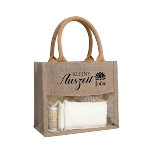 May include: A natural burlap tote bag with a clear front panel, filled with spa products. The bag has a brown handle and features the text "Kleine Auszeit Julia" in black script and a lotus flower graphic.