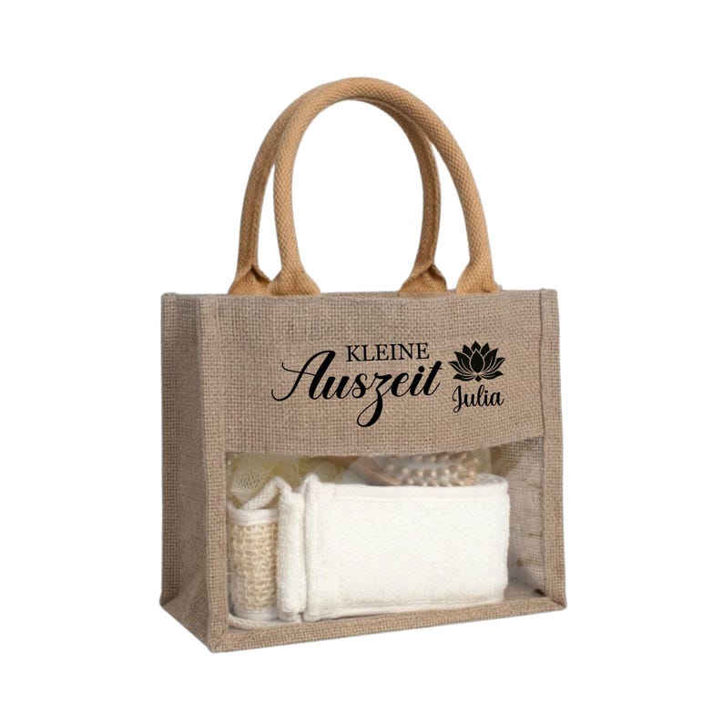 May include: A natural burlap tote bag with a clear front panel, filled with spa products. The bag has a brown handle and features the text "Kleine Auszeit Julia" in black script and a lotus flower graphic.