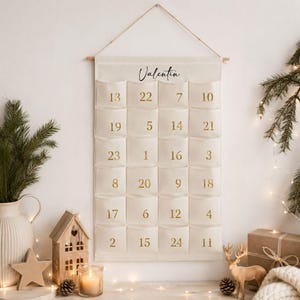 May include: A cream-coloured fabric wall hanging advent calendar with twenty-four pockets, each printed with a gold number. The name Valentin is printed in black script at the top. The calendar is suspended from a wooden rod with a string.