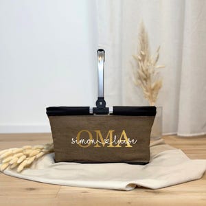 May include: A brown and black tote bag with a silver handle. The bag features the word "OMA" in gold lettering, with the names "Simon & Luisa" below. The bag is resting on a beige cloth.