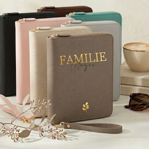 Pode incluir: Uma coleção de carteiras com zíper em várias cores, incluindo preto, rosa, bege, cinza e marrom. A carteira marrom apresenta o texto dourado "FAMILIE Mayer" e um pequeno desenho de folha dourada. Cada carteira tem uma alça de pulso.