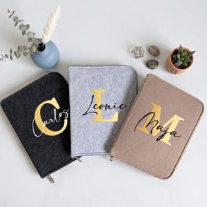 May include: Three rectangular felt zippered cases in charcoal, light gray, and beige. Each case features a large gold serif letter overlaid with a black cursive name: Carlos on the charcoal case, Leonie on the gray, and Maja on the beige case.