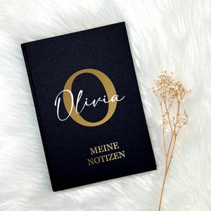 Könnte beinhalten: Ein schwarzes Notizbuch mit einem goldenen Buchstaben "O" und dem Namen "Olivia" in weißer Kursivschrift. Der Text "Meine Notizen" ist in Gold unter dem Namen gedruckt.