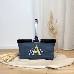 Peut inclure: Un sac fourre-tout en denim avec un dessus noir et une poignée argentée. Le sac présente la lettre "A" dorée et le nom "Annika" en écriture cursive. Le sac est posé sur un tissu beige.