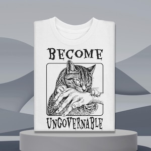 Puede incluir: Camiseta blanca con un gráfico en blanco y negro de un gato mordiendo una mano. El texto "BECOME UNGOVERNABLE" está encima y debajo del gráfico. La camiseta está doblada y expuesta sobre un pedestal gris.