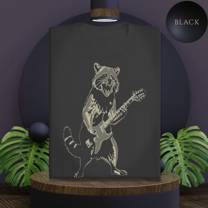 Puede incluir: Camiseta negra con una ilustración crema de un mapache tocando una guitarra eléctrica. El mapache está representado en un estilo de dibujo lineal detallado, con la boca abierta. La palabra "BLACK" se muestra en un círculo negro.