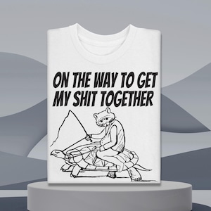 May include: White t-shirt with the text "ON THE WAY TO GET MY SHIT TOGETHER." The graphic shows a raccoon riding a turtle, holding a flag. The design is in black ink.