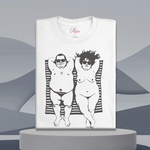 May include: White t-shirt featuring a black and white illustration of two people sunbathing on a striped towel. Both figures are wearing sunglasses and are depicted in a relaxed pose. The shirt has a crew neck and a small logo near the collar.