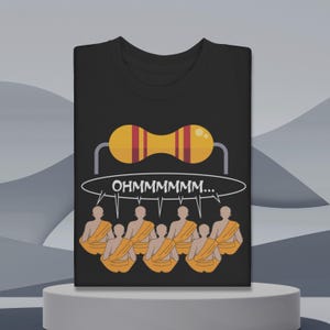 Puede incluir: Camiseta negra con un gráfico de una resistencia y el texto "OHMMMMMMM..." en un bocadillo. Debajo, ilustraciones de monjes con túnicas naranjas meditando. El diseño es una versión humorística de la electrónica y la meditación.