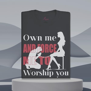 Op de afbeelding: Een donkergrijs T-shirt met een wit grafisch ontwerp. De grafiek toont een silhouet van een vrouw die op een stoel zit met een man die voor haar knielt. De tekst "Own me AND FORCE ME TO Worship you" is in wit gedrukt, met de woorden "AND FORCE ME TO" in rood.