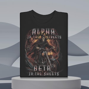 以下が含まれることがあります： 黒いTシャツ。バイクに乗った死神のグラフィック。テキスト「ALPHA IN THE STREETS」と「BETA IN THE SHEETS」が表示されています。デザインは、頭蓋骨と炎があり、ダークでエッジの効いた美学を持っています。