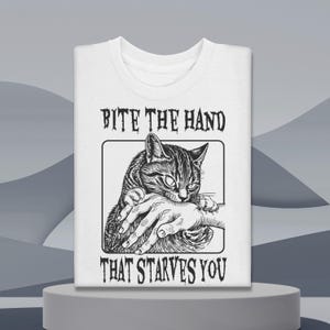 May include: White t-shirt featuring a black and white illustration of a cat biting a hand. The text "BITE THE HAND THAT STARVES YOU" is above and below the image. The shirt is folded on a gray surface.