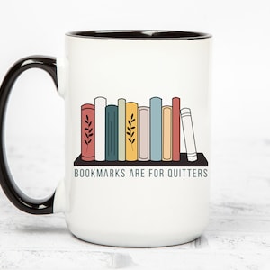 May include: White ceramic mug with a black handle and black rim. The mug has a graphic of a bookshelf with colorful books and the text "BOOKMARKS ARE FOR QUITTERS".