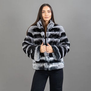 May include: A luxurious, short fur jacket in a chinchilla pattern. The jacket features a high collar and a zipper closure. The color palette includes shades of black, gray, and white, creating a striped effect. The model is wearing black pants.