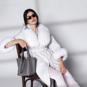 Pu&ograve; includere: Una donna indossa un cappotto bianco con collo e polsini in pelliccia, una borsa grigia e mocassini neri. Il cappotto &egrave; stretto in vita. Indossa occhiali da sole ed &egrave; seduta su una sedia di legno.