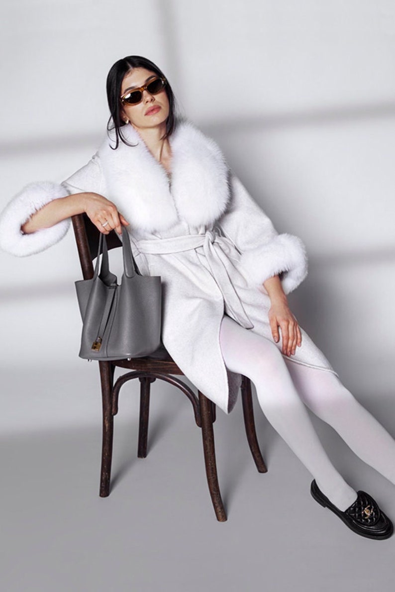 Pu&ograve; includere: Una donna indossa un cappotto bianco con collo e polsini in pelliccia, una borsa grigia e mocassini neri. Il cappotto &egrave; stretto in vita. Indossa occhiali da sole ed &egrave; seduta su una sedia di legno.