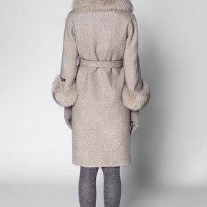 Pu&ograve; includere: Un cappotto di lana beige con collo e polsini in ecopelliccia. Il cappotto ha una cintura abbinata, maniche lunghe ed &egrave; lungo fino al ginocchio. La modella indossa collant grigi e scarpe nere. Il cappuccio &egrave; bicolore, argento e beige.