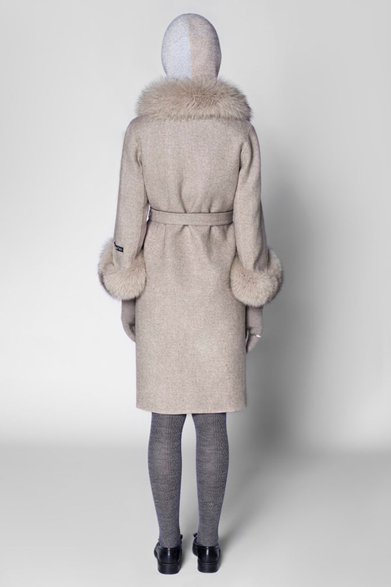 Pu&ograve; includere: Un cappotto di lana beige con collo e polsini in ecopelliccia. Il cappotto ha una cintura abbinata, maniche lunghe ed &egrave; lungo fino al ginocchio. La modella indossa collant grigi e scarpe nere. Il cappuccio &egrave; bicolore, argento e beige.