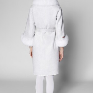 Pu&ograve; includere: Un cappotto grigio chiaro con collo e polsini in pelliccia bianca. Il cappotto ha una cintura abbinata ed &egrave; lungo fino al ginocchio. La persona indossa collant bianchi e scarpe nere. Un classico cappotto invernale.