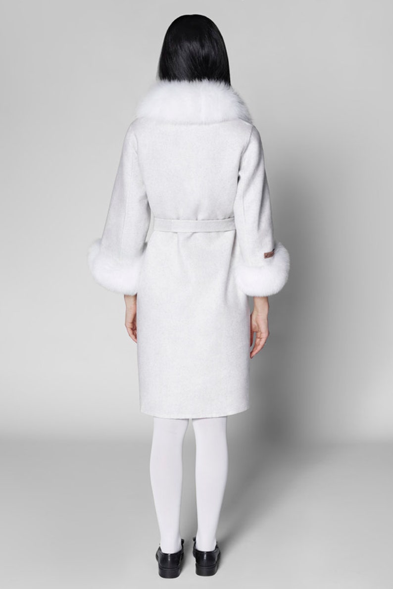 Pu&ograve; includere: Un cappotto grigio chiaro con collo e polsini in pelliccia bianca. Il cappotto ha una cintura abbinata ed &egrave; lungo fino al ginocchio. La persona indossa collant bianchi e scarpe nere. Un classico cappotto invernale.