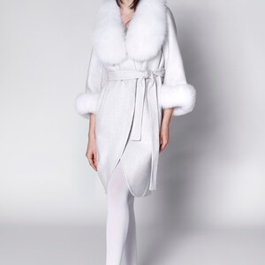 Pu&ograve; includere: Un cappotto avvolgente grigio chiaro con collo e polsini in pelliccia bianca. Il cappotto &egrave; legato in vita e indossato con collant bianchi e mocassini neri. Il design del cappotto &egrave; elegante e sofisticato.