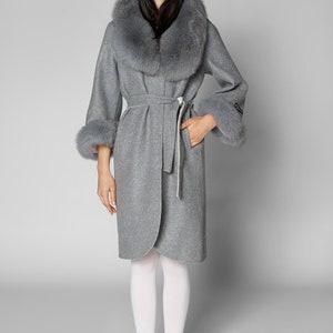Pu&ograve; includere: Un cappotto grigio in lana con collo e polsini in ecopelliccia. Il cappotto ha una cintura in vita e maniche a tre quarti. La modella indossa collant bianchi e scarpe bianche. Un capo di abbigliamento elegante.