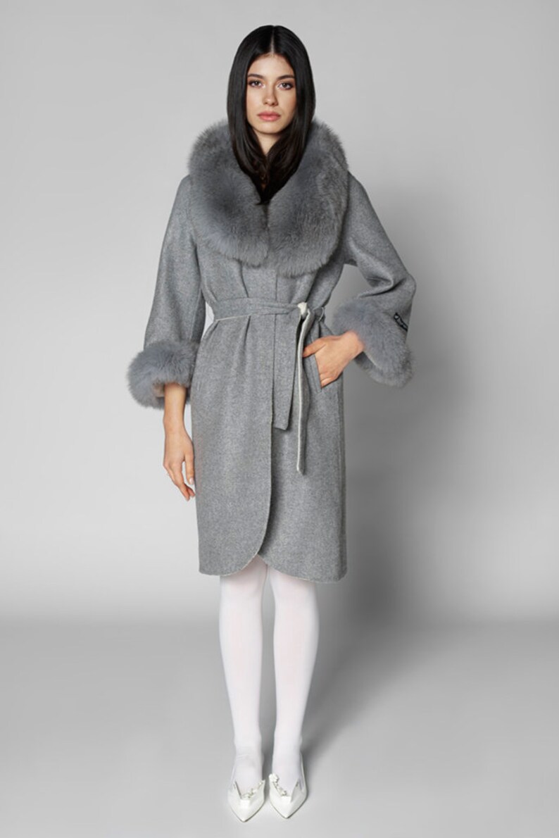 Pu&ograve; includere: Un cappotto grigio in lana con collo e polsini in ecopelliccia. Il cappotto ha una cintura in vita e maniche a tre quarti. La modella indossa collant bianchi e scarpe bianche. Un capo di abbigliamento elegante.