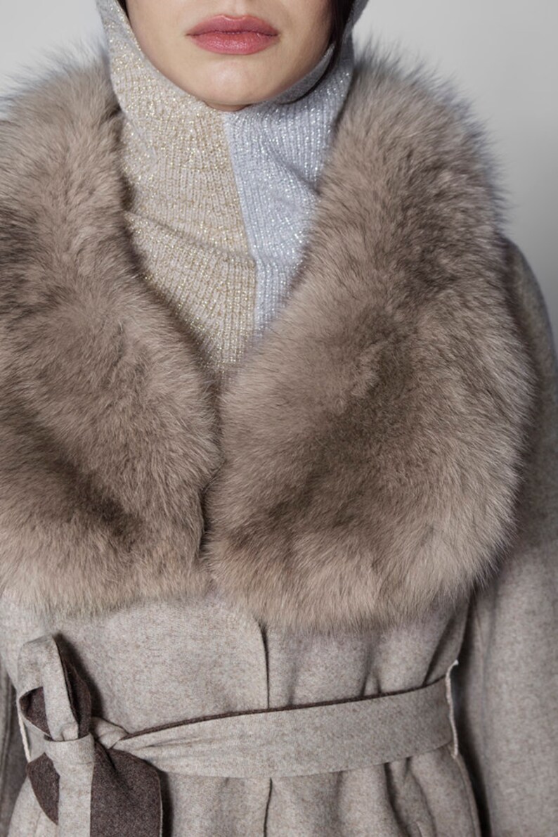 Pu&ograve; includere: Cappotto grigio chiaro con un ampio colletto in pelliccia soffice marrone chiaro. Il cappotto ha una cintura abbinata annodata a fiocco. Il colletto incornicia un dolcevita a due toni in argento e oro.