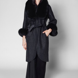 Pu&ograve; includere: Un cappotto grigio scuro con collo e polsini in pelliccia nera. Il cappotto ha una cintura in vita e un design lungo fino al ginocchio. La modella indossa pantaloni neri. Un elegante capo di abbigliamento.