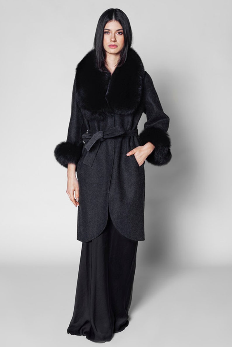 Pu&ograve; includere: Un cappotto grigio scuro con collo e polsini in pelliccia nera. Il cappotto ha una cintura in vita e un design lungo fino al ginocchio. La modella indossa pantaloni neri. Un elegante capo di abbigliamento.