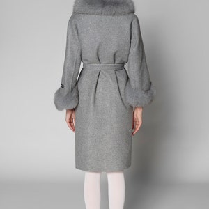 Pu&ograve; includere: Un cappotto grigio in lana con collo e polsini in ecopelliccia. Il cappotto presenta una cintura in vita e un orlo al ginocchio. La modella indossa collant bianchi e scarpe bianche. Design classico ed elegante.