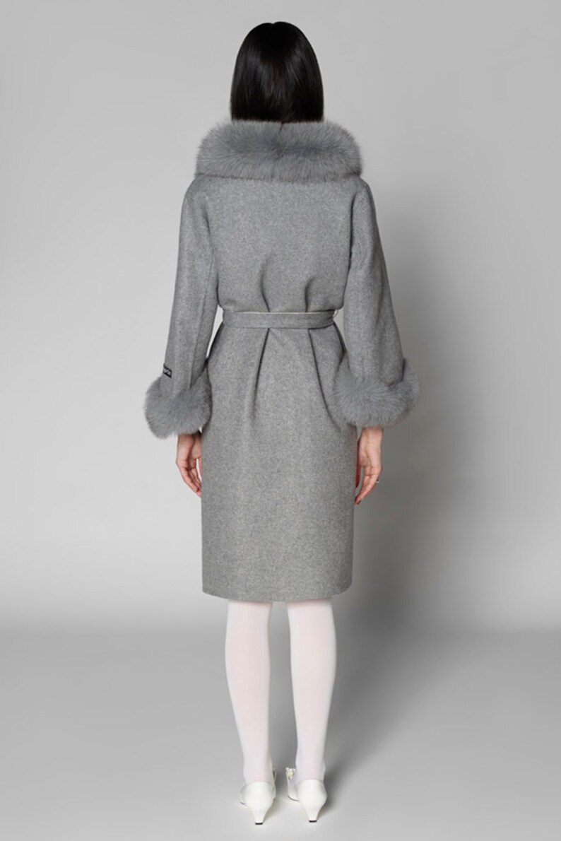 Pu&ograve; includere: Un cappotto grigio in lana con collo e polsini in ecopelliccia. Il cappotto presenta una cintura in vita e un orlo al ginocchio. La modella indossa collant bianchi e scarpe bianche. Design classico ed elegante.