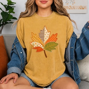 May include: A mustard yellow crew neck t-shirt featuring a large central graphic of a maple leaf. The leaf is divided into sections with various patterns including brown and white gingham, white polka dots, and floral prints in orange and green.