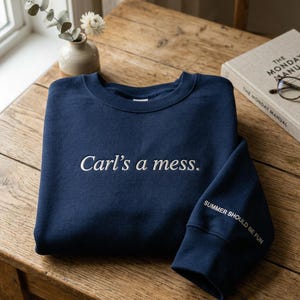 May include: Navy blue sweatshirt with the phrase "Carl's a mess." embroidered in white on the front. The sleeve cuff reads "SUMMER SHOULD BE FUN." The sweatshirt is folded on a wooden table.
