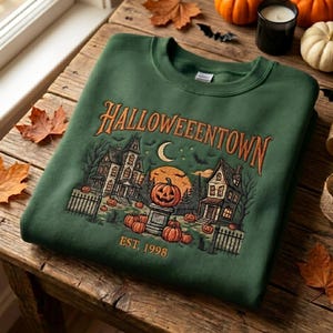 Pode incluir: Uma camisola de gola redonda verde-floresta com uma impressão gráfica com o texto HALLOWEENTOWN EST. 1998. A ilustração mostra duas casas, uma lua crescente, morcegos e várias abóboras dispostas num pedestal e no chão.