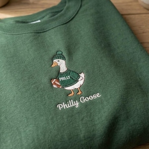 May include: A forest green crewneck sweatshirt featuring an embroidered graphic of a white goose wearing a green knit hat and a green jumper with the word PHILLY printed on it. The goose holds a brown football. The text Philly Goose is below.
