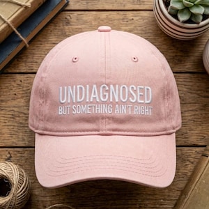 Può includere: Un cappellino da baseball rosa chiaro con testo ricamato in bianco sulla parte anteriore che recita UNDIAGNOSED BUT SOMETHING AIN'T RIGHT. Il cappellino presenta una corona strutturata, una visiera curva con cuciture visibili e un bottone centrale.