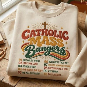 May include: A beige crewneck sweatshirt featuring the text CATHOLIC MASS BANGERS in orange, yellow, and green, topped with a cross. Below, a numbered list of ten song titles is printed in orange and green, including On Eagle's Wings.