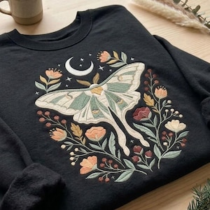 May include: A black long-sleeve sweatshirt featuring an embroidered design of a large moth with pale green wings, centered beneath a white crescent moon and stars, surrounded by a floral arrangement of orange, red, and green flowers.