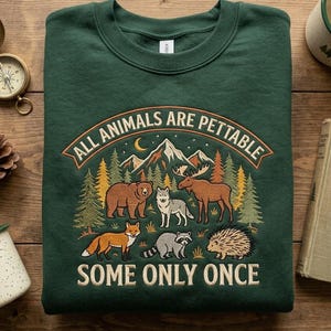May include: A dark green crewneck sweatshirt featuring a graphic of a bear, wolf, moose, fox, raccoon, and hedgehog in a forest with mountains. The text reads ALL ANIMALS ARE PETTABLE SOME ONLY ONCE in a tan and orange arched design.