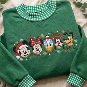May include: A forest green sweatshirt featuring a green and white gingham collar and cuffs. The center displays embroidered faces of Mickey Mouse, Minnie Mouse, Donald Duck, Goofy, and Pluto wearing holiday hats, surrounded by snowflakes.