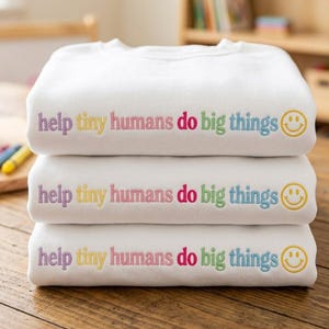 Puede incluir: Una pila de tres sudaderas blancas, cada una con el texto bordado "help tiny humans do big things" en letras multicolores, seguido de un icono de cara sonriente amarilla bordado en el lado derecho del área del pecho.