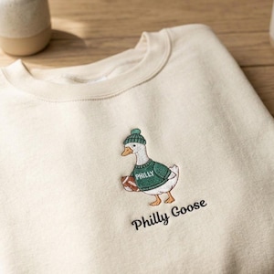 May include: A cream-colored crewneck sweatshirt featuring an embroidered graphic of a white goose wearing a green knit hat and a green sweater with the word PHILLY printed on it, holding a brown football. The text Philly Goose is embroidered below.