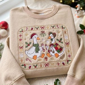 May include: A beige crewneck sweatshirt featuring a central embroidered rectangular panel. The design depicts two white geese wearing holiday accessories, surrounded by a border of small Christmas icons including candy canes, holly, and bows.