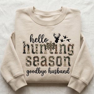 Könnte beinhalten: Ein beigefarbenes Sweatshirt mit Rundhalsausschnitt, schwarzem Text "hello" und "goodbye husband" sowie dem Schriftzug "hunting season" in Camouflage-Muster. Das Design zeigt eine schwarze Hirschsilhouette, eine kleine Schleife und drei Enten.