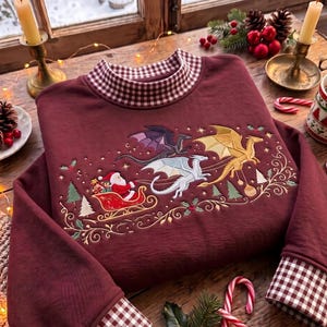 May include: A maroon sweatshirt featuring a high collar and cuffs with a brown and white gingham pattern. The front displays an embroidered scene of Santa Claus in a red sleigh pulled by three dragons in purple, white, and gold colors.