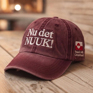 May include: A maroon baseball cap with white embroidered text that reads "Nu det NUUK!" on the front. The cap has a small patch with a Greenland flag and the words "Stand with Greenland". The cap is sitting on a wooden surface.