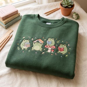 Pu&ograve; includere: Una felpa girocollo verde scuro con un ricamo raffigurante quattro rane verde chiaro che interagiscono con libri e un fungo. Le rane sono raffigurate mentre leggono, reggono pile di libri e sono sedute su un fungo rosso a pois bianchi.