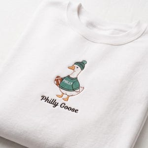 May include: A white crewneck sweatshirt featuring an embroidered design of a goose wearing a green knit hat and a green sweater with the word PHILLY printed on it. The goose holds a brown football, and the text Philly Goose is embroidered below.