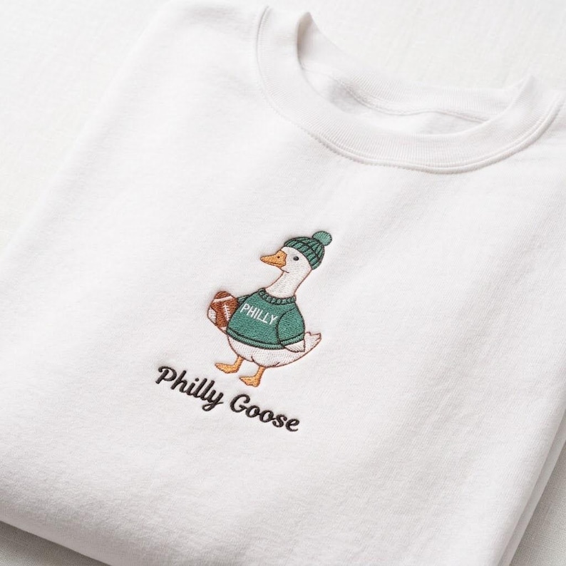 May include: A white crewneck sweatshirt featuring an embroidered design of a goose wearing a green knit hat and a green sweater with the word PHILLY printed on it. The goose holds a brown football, and the text Philly Goose is embroidered below.